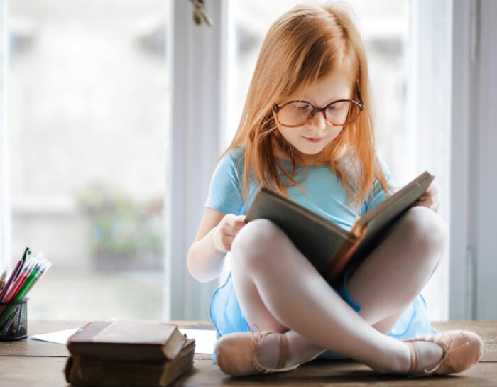 Jeune fille en tailleur sur une table lit un livre. Elle porte des lunettes trop grandes pour elle