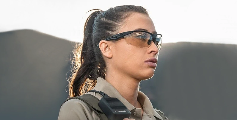 Une femme porte des lunettes de balistique de la marque Bollé Safety. Elle est dehors et porte des vêtements militaires. Elle a l'air très concentrée