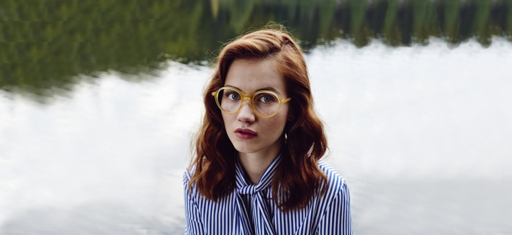 Une jeune femme rousse porte des lunettes de la marque Massada. Elle est eu bord d'un point d'eau et d'une forêt